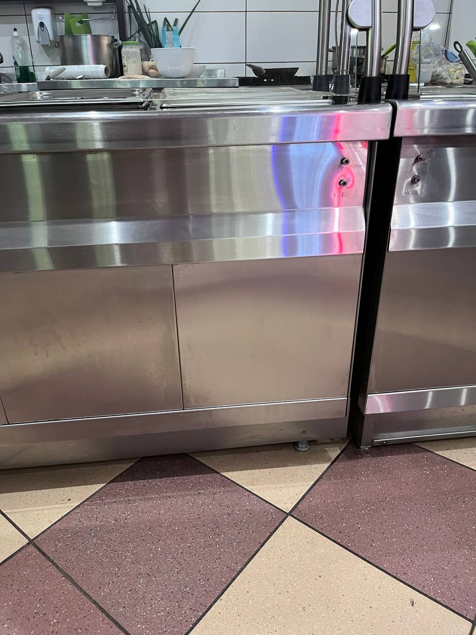 Błyszcząca, metalowa szafka w kuchni z widocznymi elementami wyposażenia na blacie i wzorzystą podłogą w odcieniach brązu i beżu.