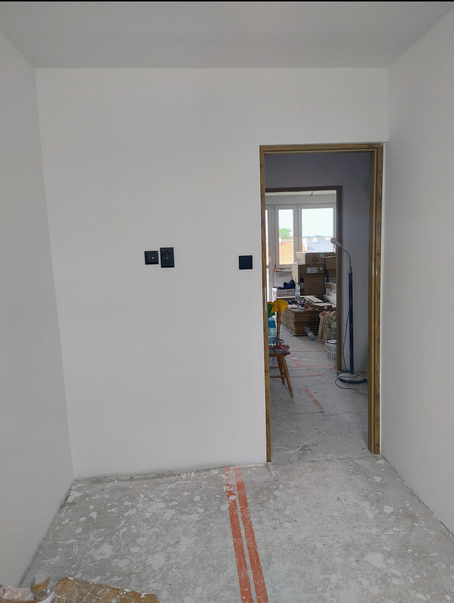 Wnętrze w trakcie wykończenia: świeżo pomalowane ściany z zamontowanymi gniazdkami, surowa podłoga z widocznymi śladami prac, drewniana ościeżnica drzwiowa.