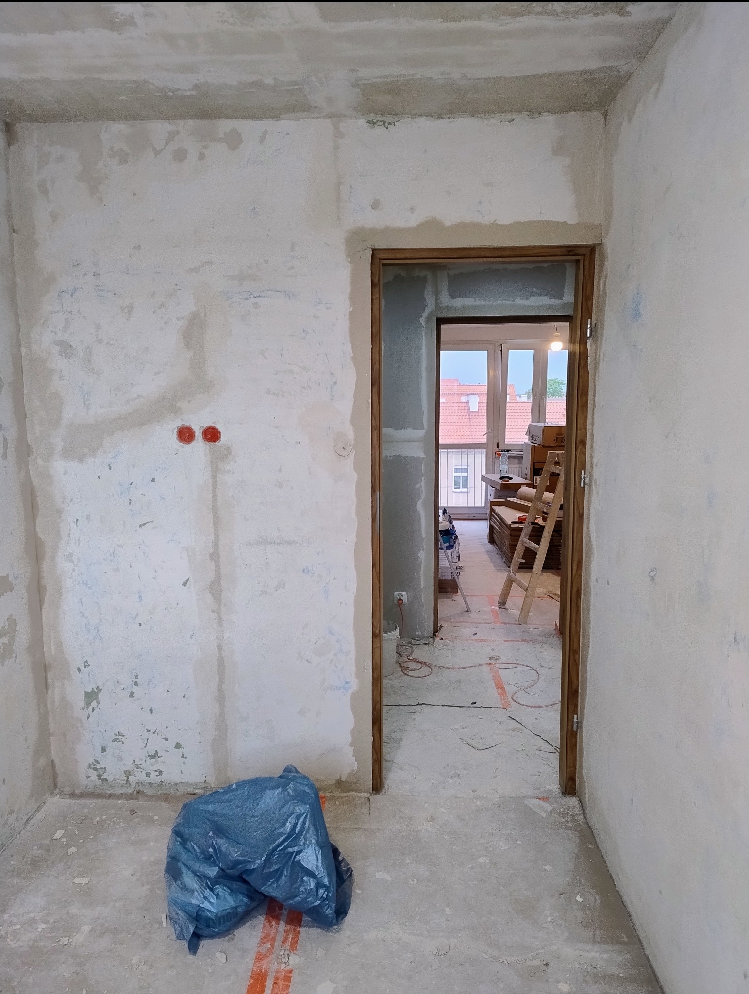 Wnętrze w trakcie remontu w Mikołajkach: surowe ściany, widoczne instalacje elektryczne, worek z odpadami budowlanymi i otwarte przejście do kolejnego pomieszczenia.