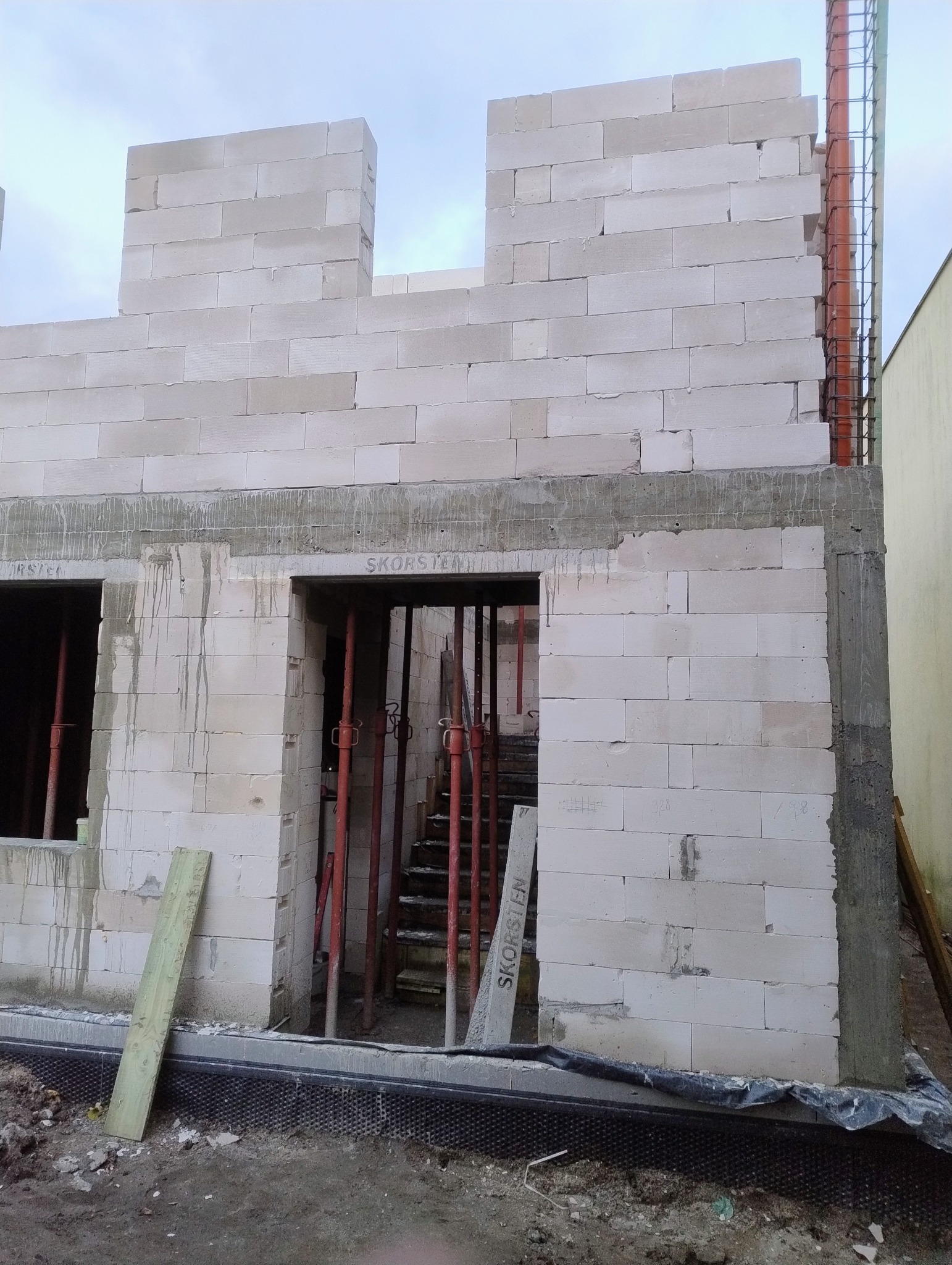 Wznoszenie ścian parteru domu z betonu komórkowego. Widoczne otwory okienne i drzwiowe, podpory szalunkowe oraz zbrojenie pionowe. Etap budowy w toku.