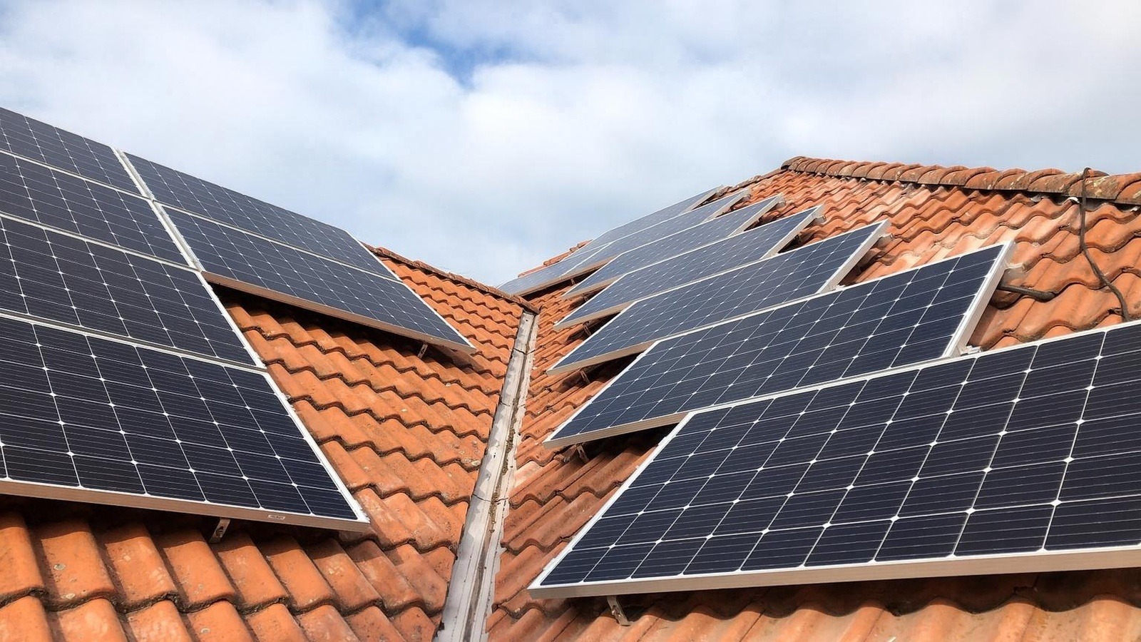 Panele słoneczne zamontowane na dachu z czerwonej dachówki, widok z dołu pod kątem. Widoczne mocowania paneli i fragment nieba z chmurami.