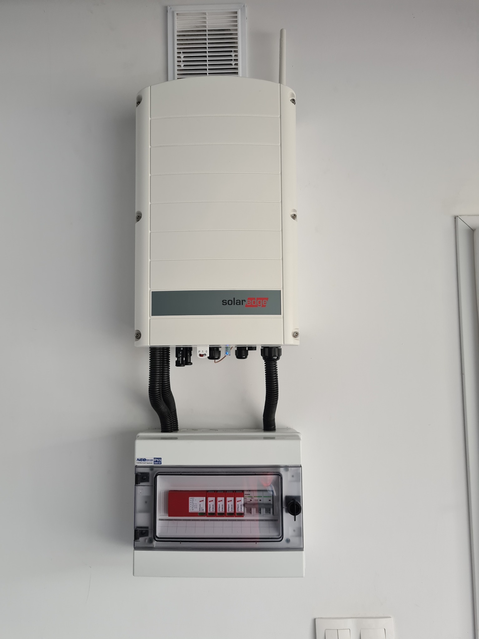 Biały inwerter SolarEdge z czarnymi przewodami i skrzynką bezpiecznikową na białej ścianie. Widoczny fragment wentylacji i gniazdka elektrycznego.