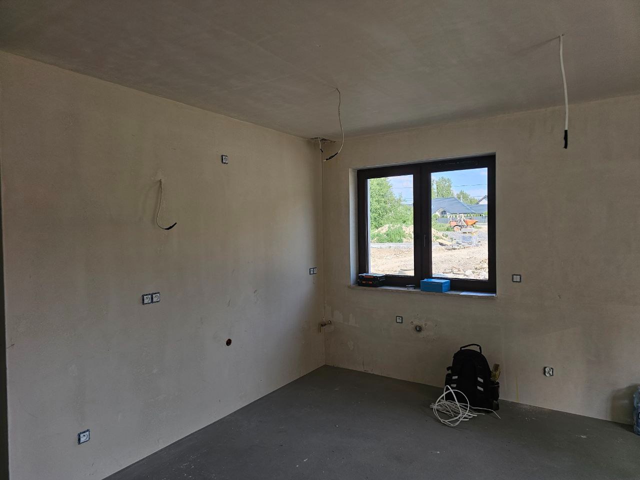 Surowy pokój z widocznymi puszkami elektrycznymi w ścianach, kable zwisające z sufitu i torba narzędziowa na podłodze. Widok z okna na zewnątrz.