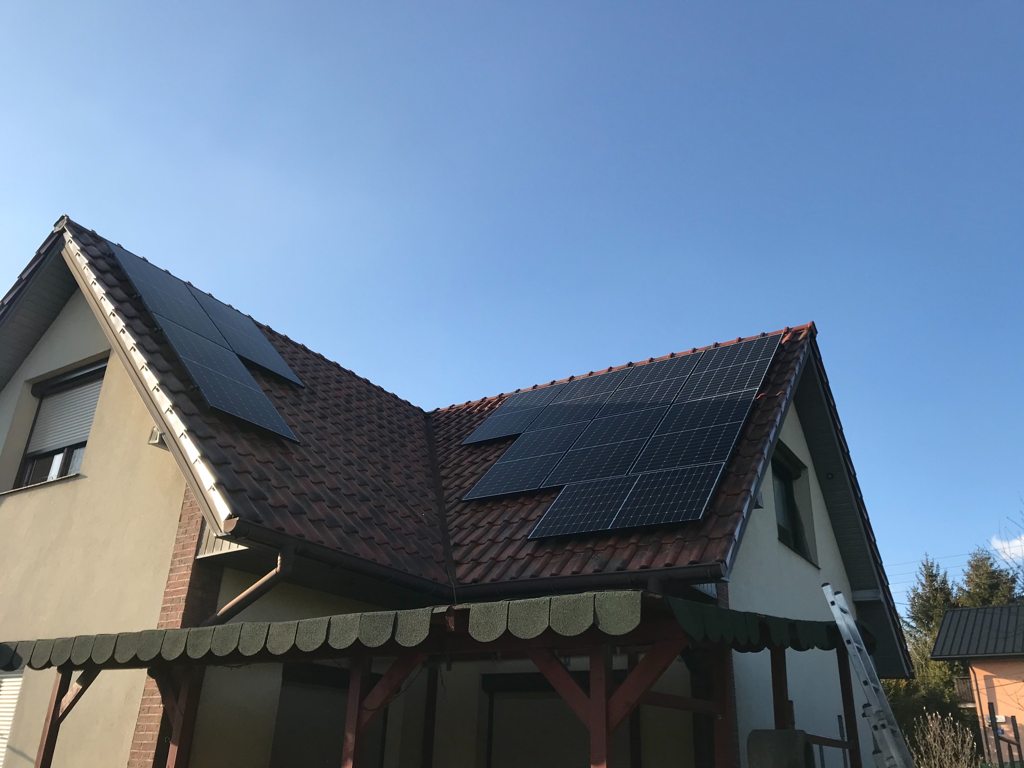 Dom z panelami słonecznymi na dachu z czerwonej dachówki, widziany z dołu pod kątem. Aluminiowa drabina oparta o ścianę. Błękitne niebo w tle.