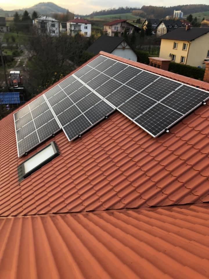 Panele słoneczne na dachu z dachówki ceramicznej, widok z góry, w tle zabudowa jednorodzinna i zalesione wzgórza, pochmurne niebo.