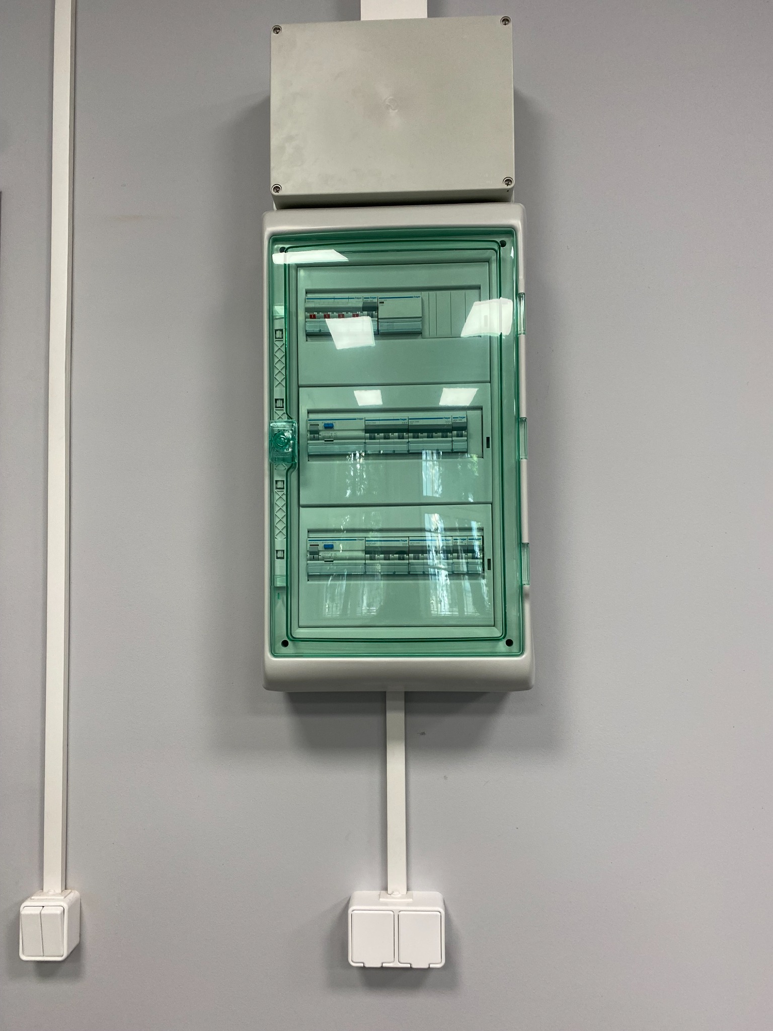 Rozdzielnica elektryczna z zieloną osłoną, zamontowana na szarej ścianie, widoczne kable. Usługi elektryczne w Krakowie.