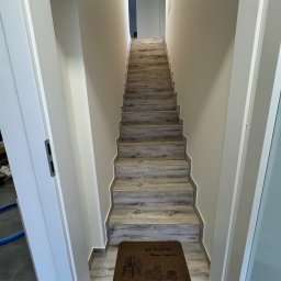 Ivan Shubin - Schody wykończone panelami imitującymi drewno, prowadzące w górę, z wycieraczką 'at home' u dołu. Ściany pomalowane na jasny kolor, widoczne białe futryny drzwi.