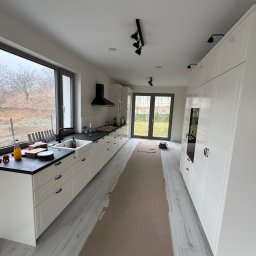 Ivan Shubin - Jasna kuchnia z białymi szafkami i czarnym blatem, widok na okno i drzwi balkonowe. Podłoga wyłożona panelami imitującymi drewno. Oświetlenie punktowe.