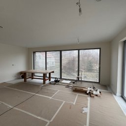 Ivan Shubin - Przygotowanie do remontu: zabezpieczona podłoga kartonem, stół roboczy, okna z żaluzjami. Widoczne narzędzia i materiały budowlane. Wnętrze w trakcie prac wykończeniowych.