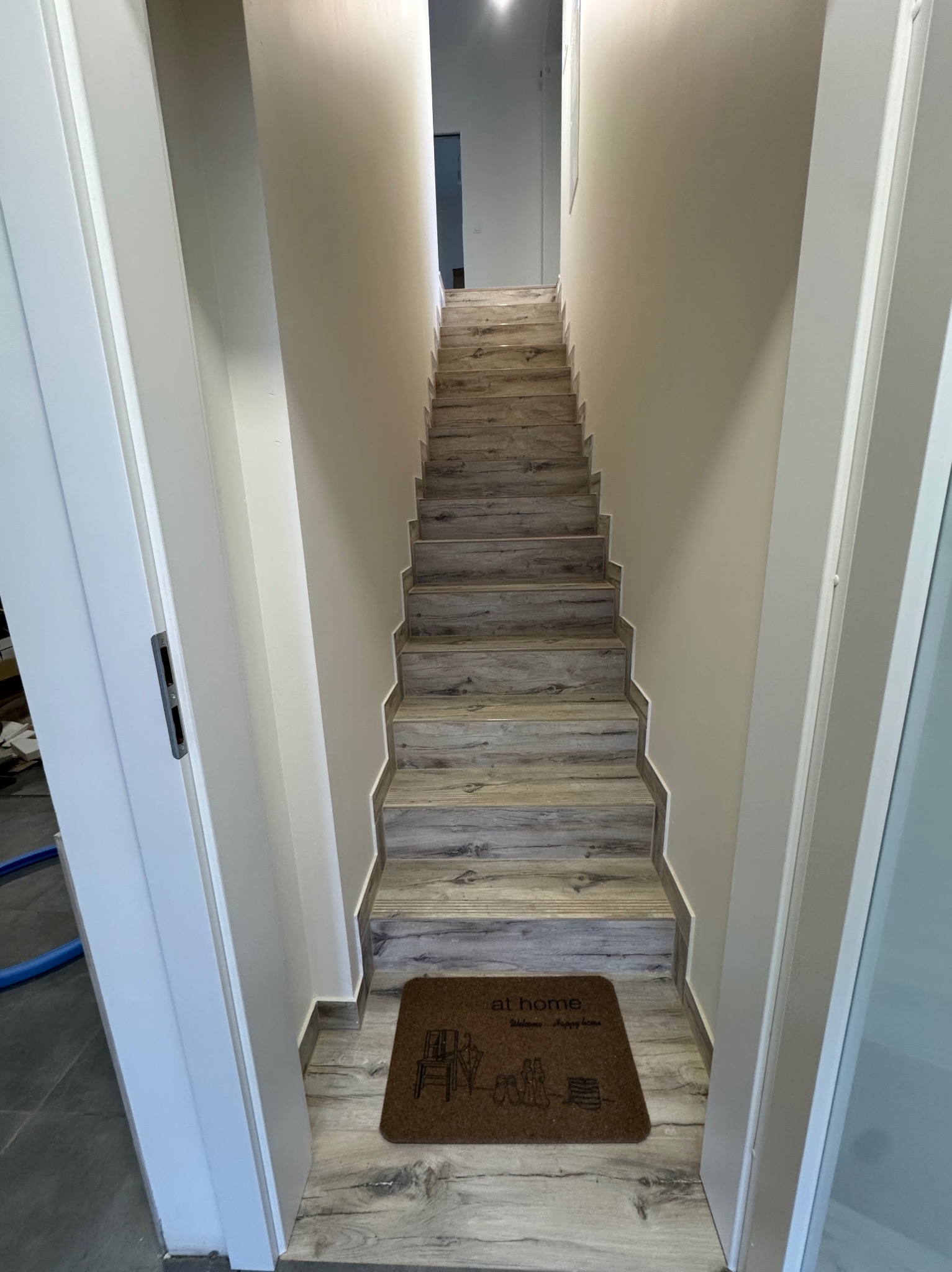 Schody wykończone panelami imitującymi drewno, prowadzące w górę, z wycieraczką 'at home' u dołu. Ściany pomalowane na jasny kolor, widoczne białe futryny drzwi.