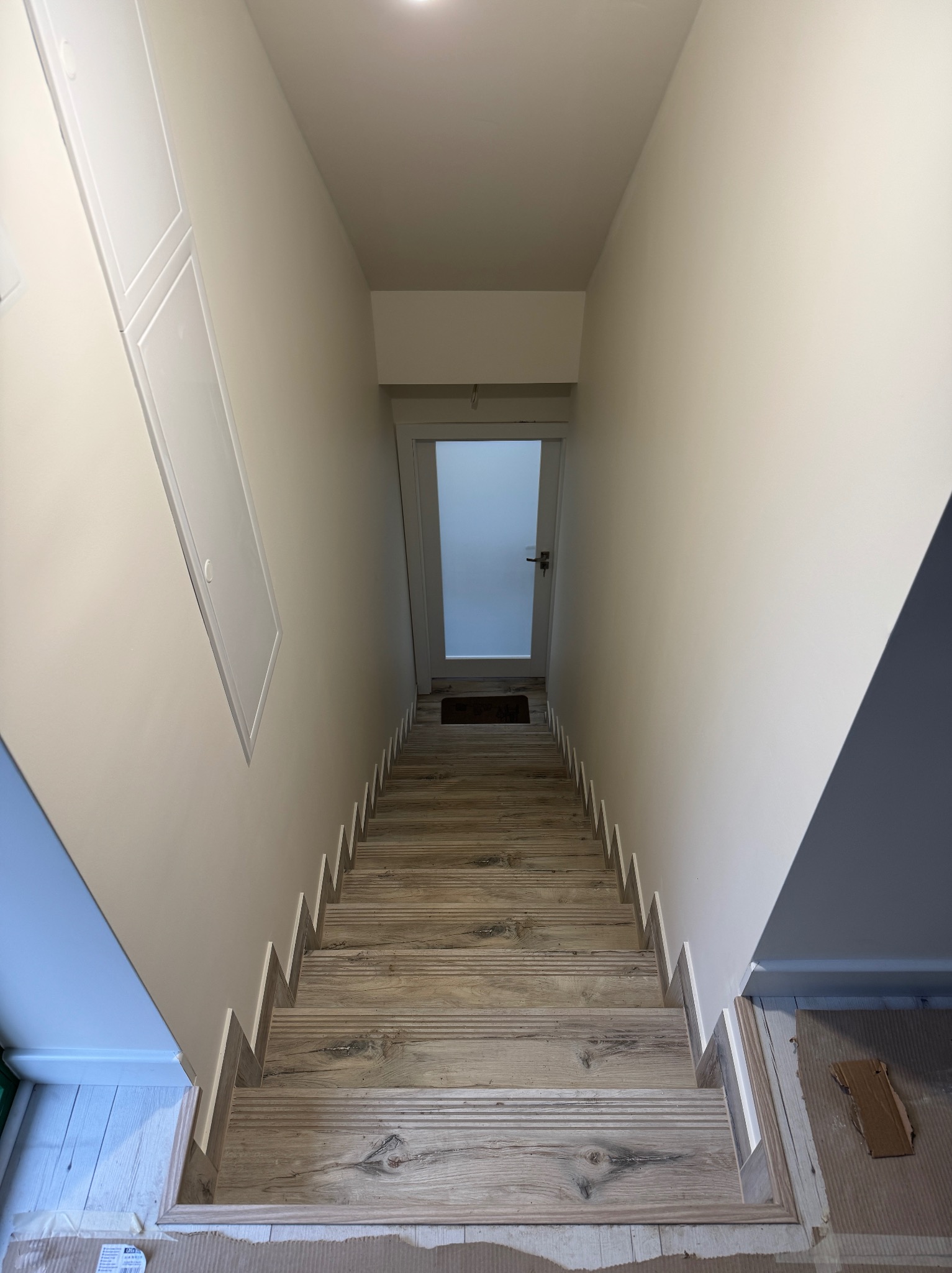 Wykończone schody z drewnopodobnymi płytkami, prowadzące do drzwi. Widoczne listwy przypodłogowe i fragment sufitu z oświetleniem. Ujęcie z góry.