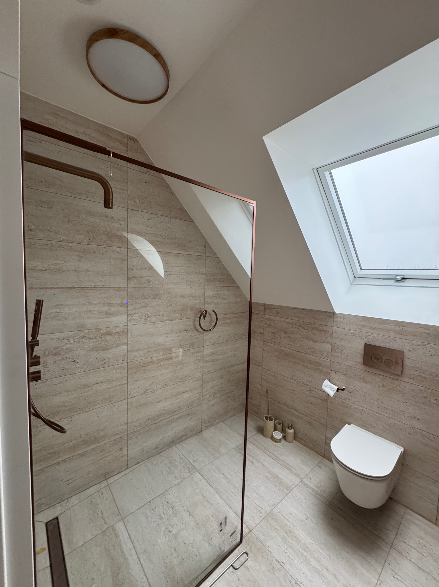 Nowoczesna łazienka na poddaszu z kabiną prysznicową typu walk-in, wykończona beżowymi płytkami imitującymi trawertyn i oświetlona okrągłą lampą sufitową.