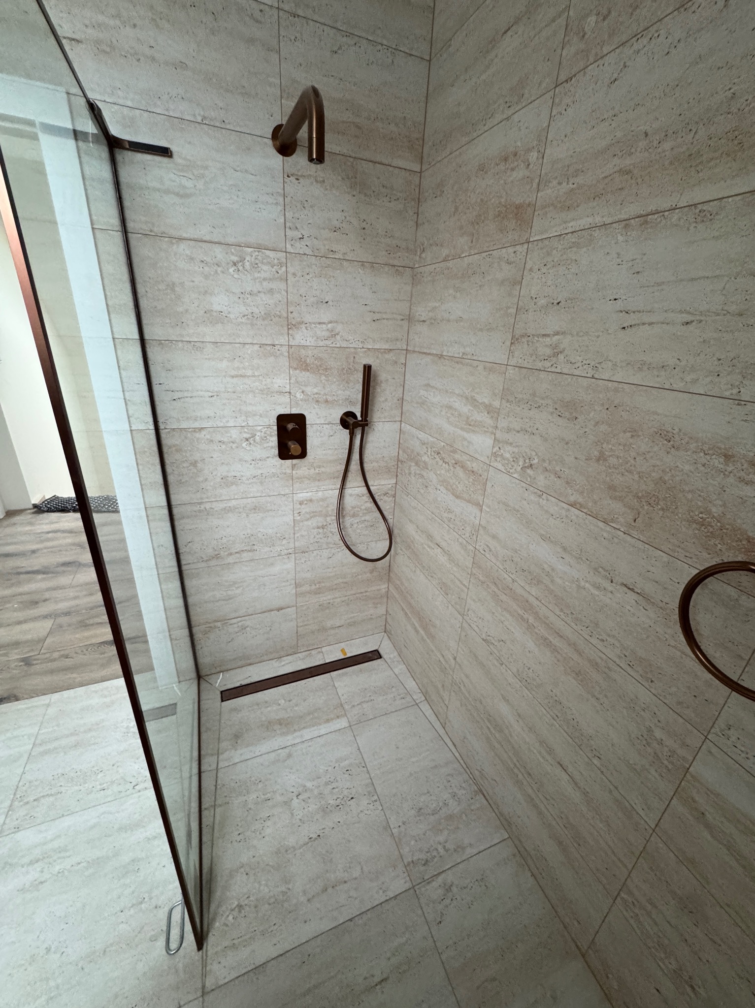 Nowoczesna kabina prysznicowa z beżowymi płytkami imitującymi kamień, armatura w kolorze miedzi, szklana ściana kabiny. Minimalistyczny design, eleganckie wykończenie.