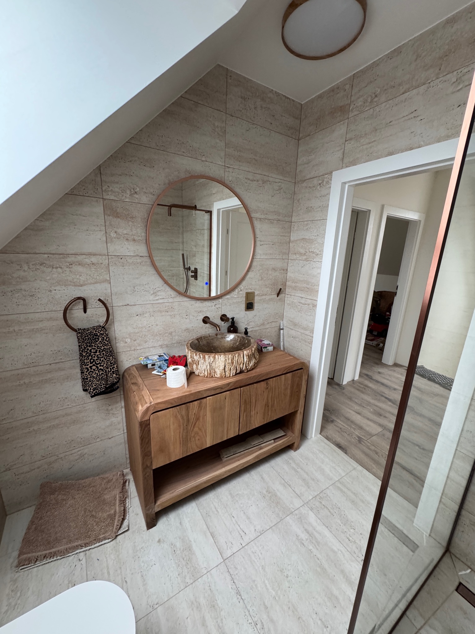 Nowoczesna łazienka na poddaszu z drewnianą szafką pod umywalkę, okrągłym lustrem w miedzianej ramie i beżowymi płytkami. Widoczny fragment toalety i drzwi.