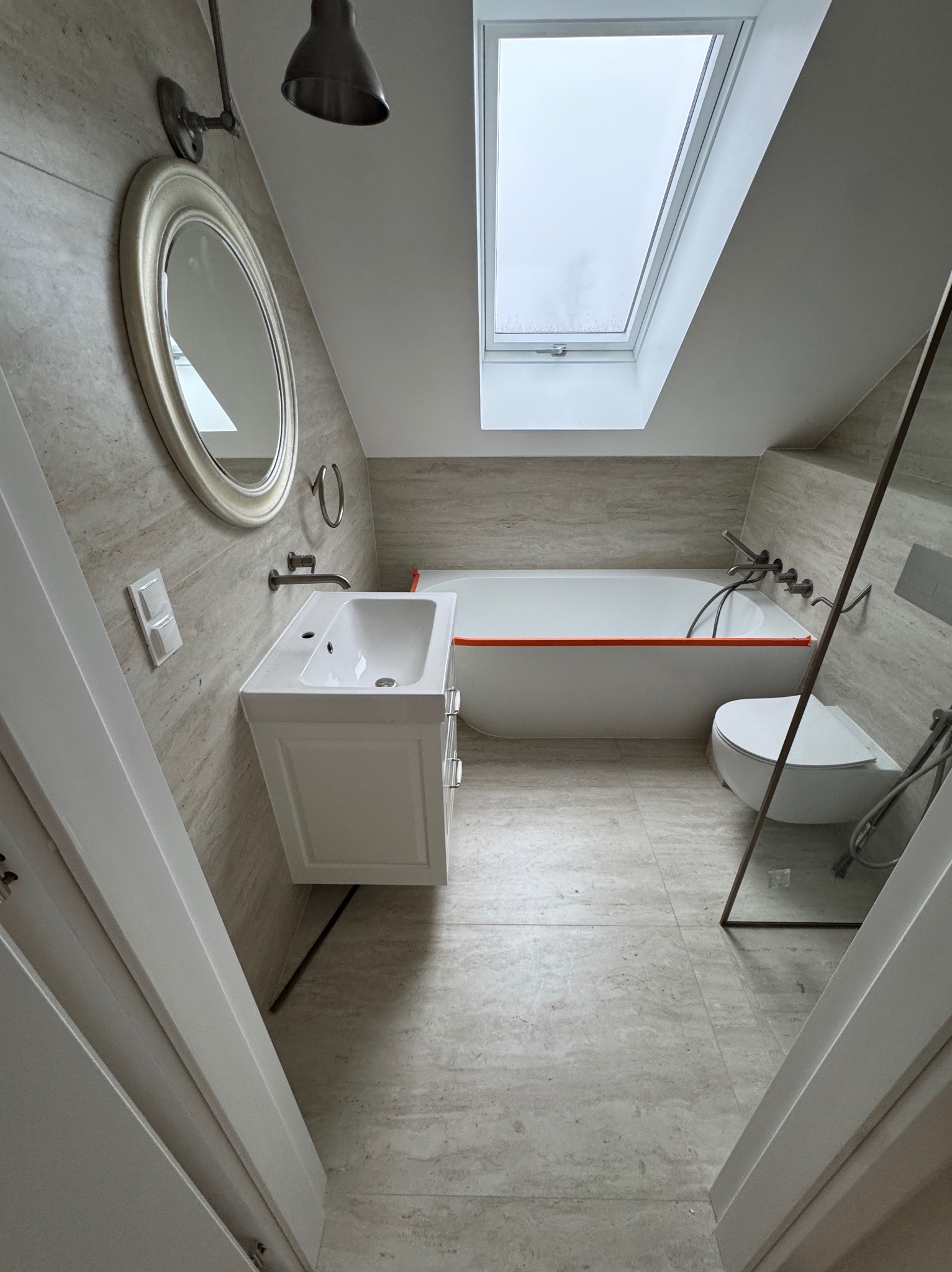 Jasna łazienka na poddaszu z oknem dachowym. Umywalka z szafką, wanna z pomarańczowym akcentem, toaleta i lustro. Beżowe płytki na ścianach i podłodze.