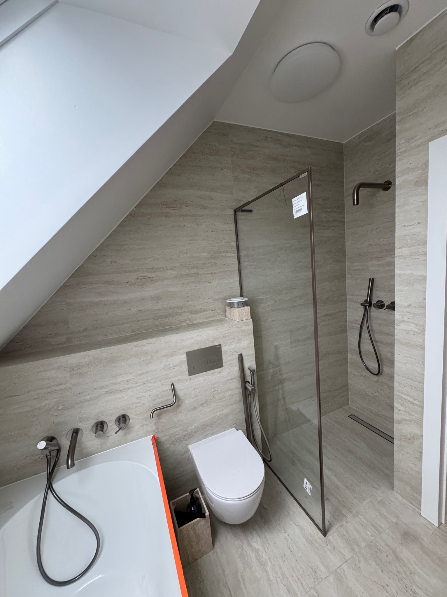 Nowoczesna łazienka na poddaszu z beżowymi płytkami imitującymi trawertyn, kabiną prysznicową typu walk-in i minimalistyczną armaturą w kolorze grafitowym.