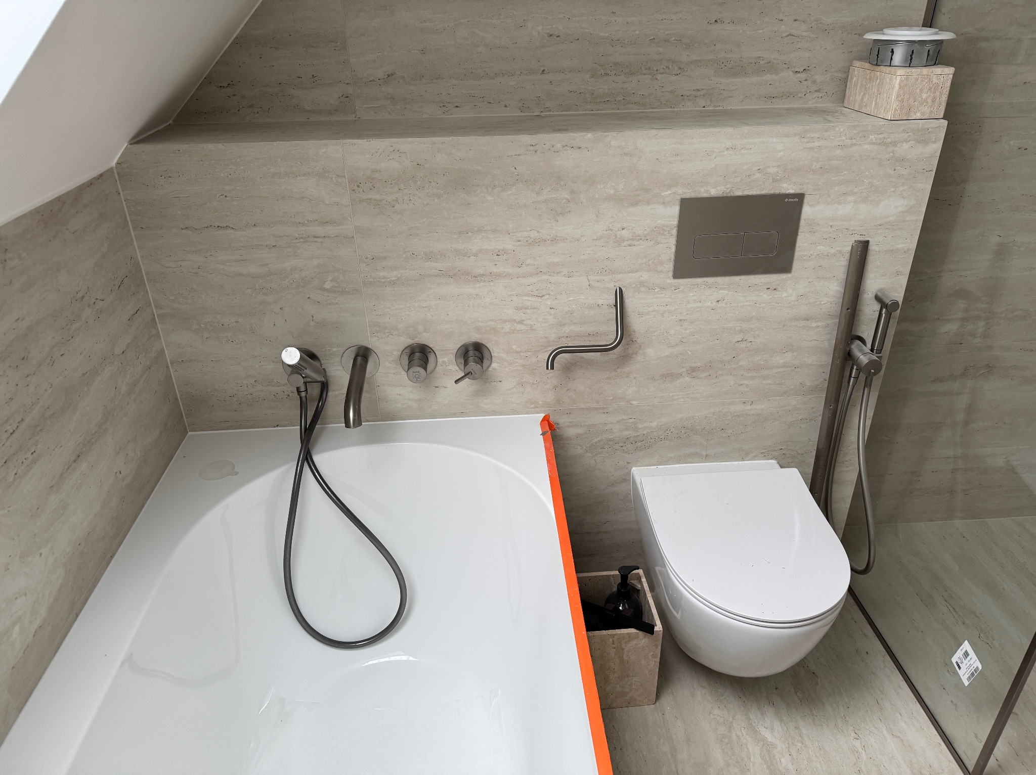 Wykończona łazienka na poddaszu we Wrocławiu. Beżowe płytki imitujące kamień, biała wanna z pomarańczowym zabezpieczeniem, toaleta podtynkowa i szklana kabina.