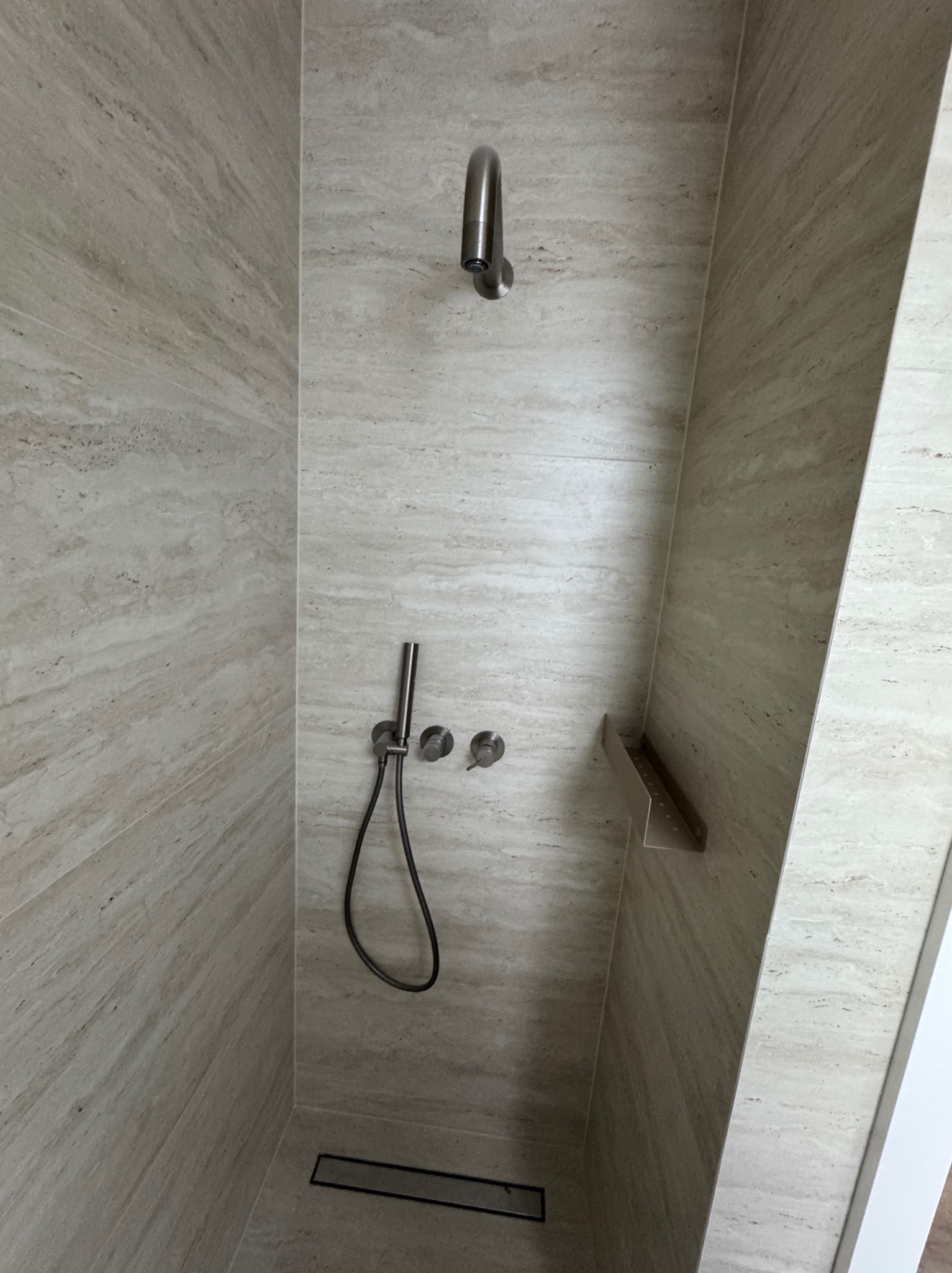 Beżowa kabina prysznicowa z deszczownicą i odpływem liniowym, wykończona płytkami imitującymi trawertyn. Minimalistyczny design z półką wnękową.