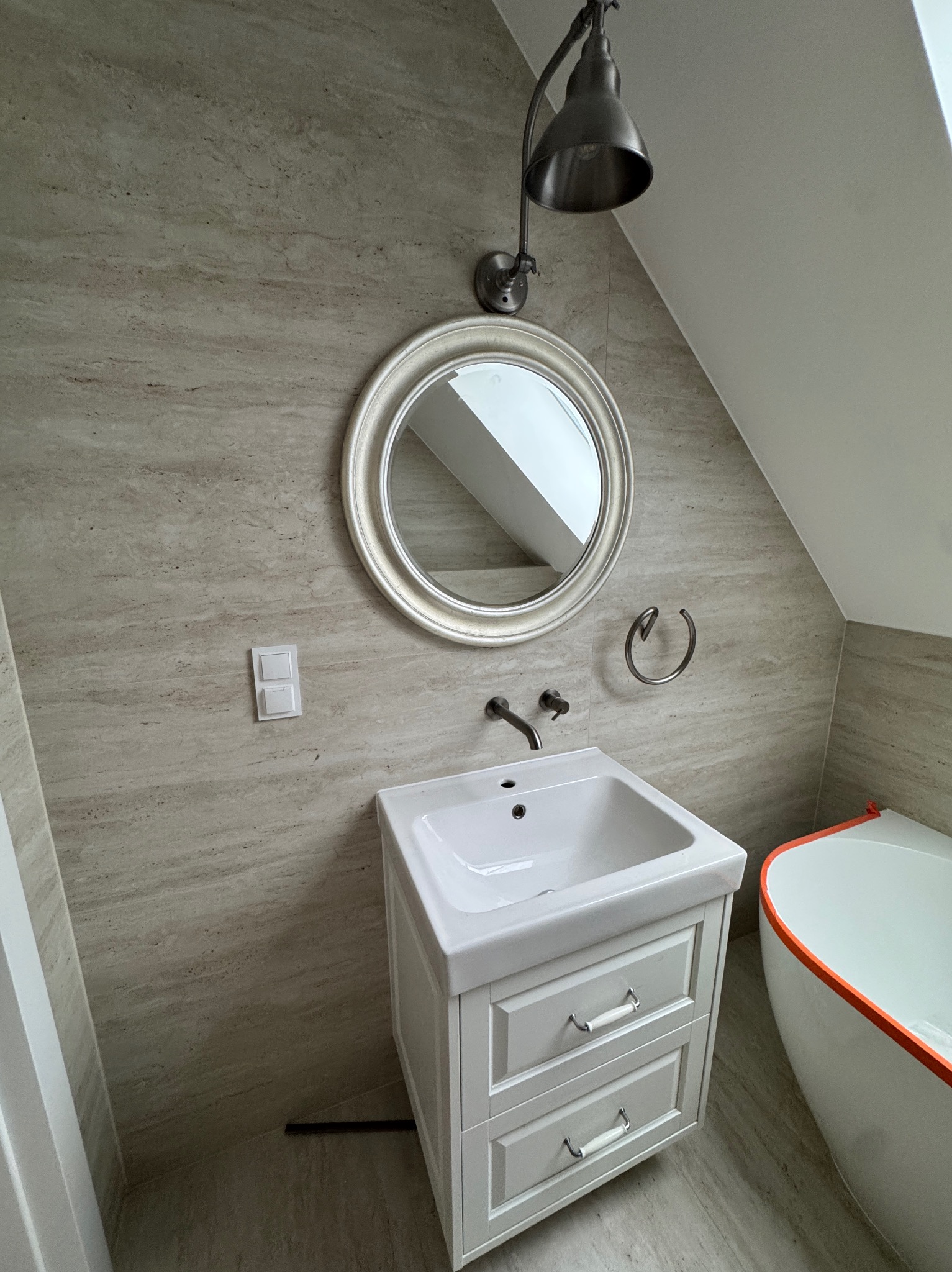 Jasna łazienka na poddaszu: umywalka z szafką, okrągłe lustro w srebrnej ramie, beżowe płytki imitujące kamień, nowoczesna lampa i fragment wanny.