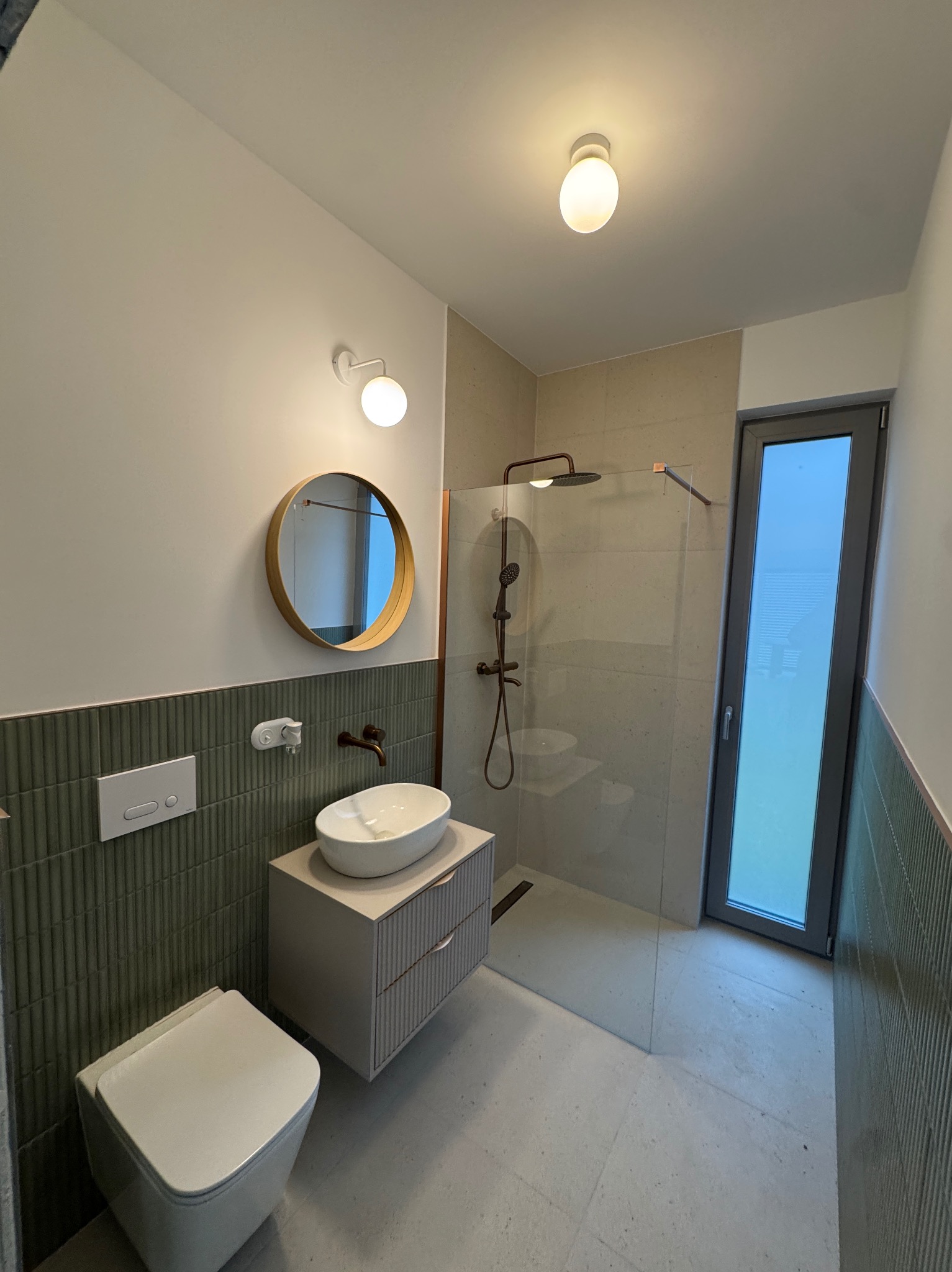 Nowoczesna łazienka po remoncie: zielone płytki, okrągłe lustro, biała umywalka nablatowa i kabina prysznicowa ze szklaną ścianką. Minimalistyczny design.