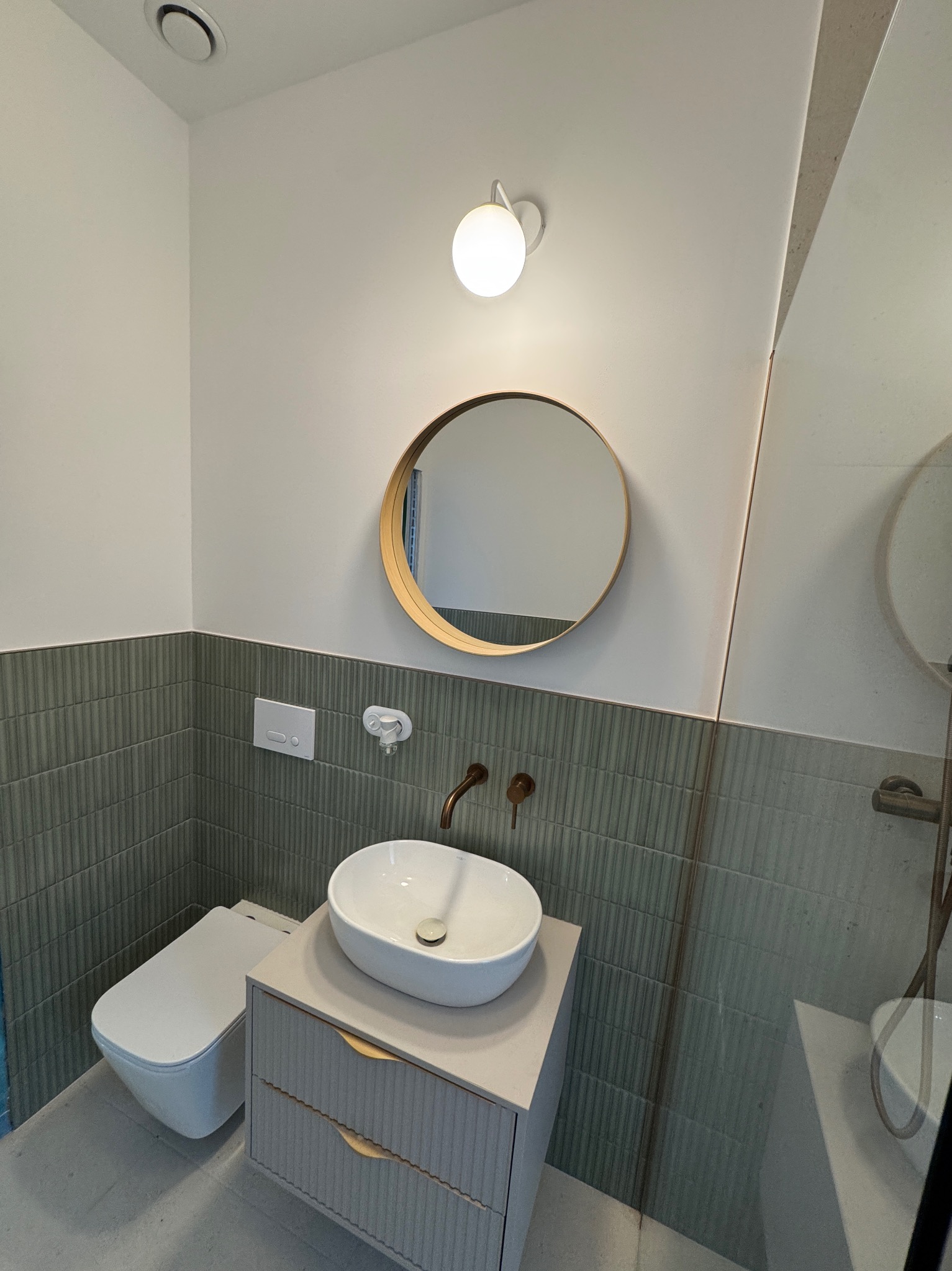 Nowoczesna łazienka z umywalką nablatową, okrągłym lustrem w złotej ramie i szarozielonymi płytkami. Minimalistyczny design, ciepłe oświetlenie.