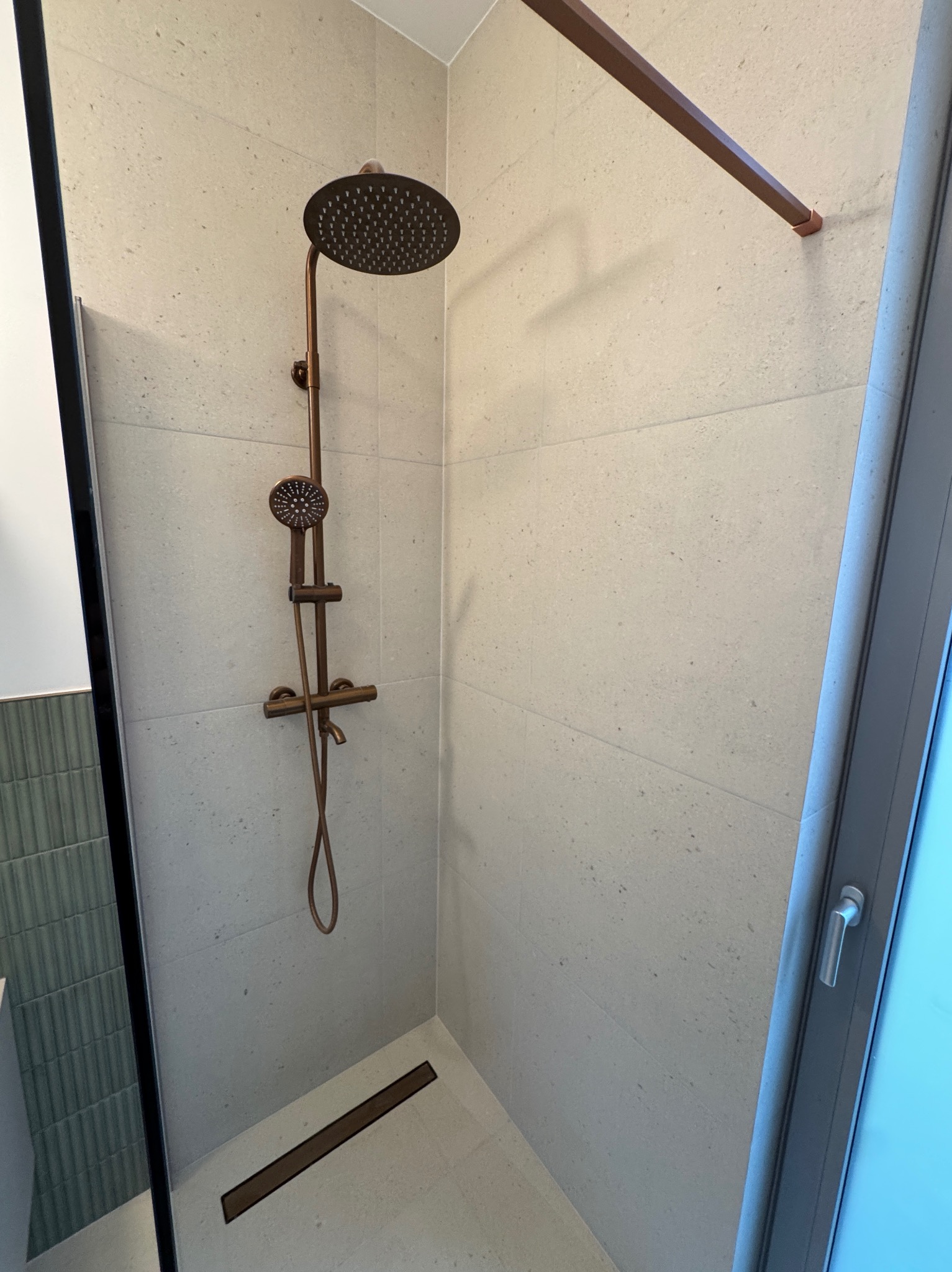 Nowoczesna kabina prysznicowa z beżowymi płytkami, deszczownicą i odpływem liniowym w kolorze miedzi. Minimalistyczny design, szklane drzwi.