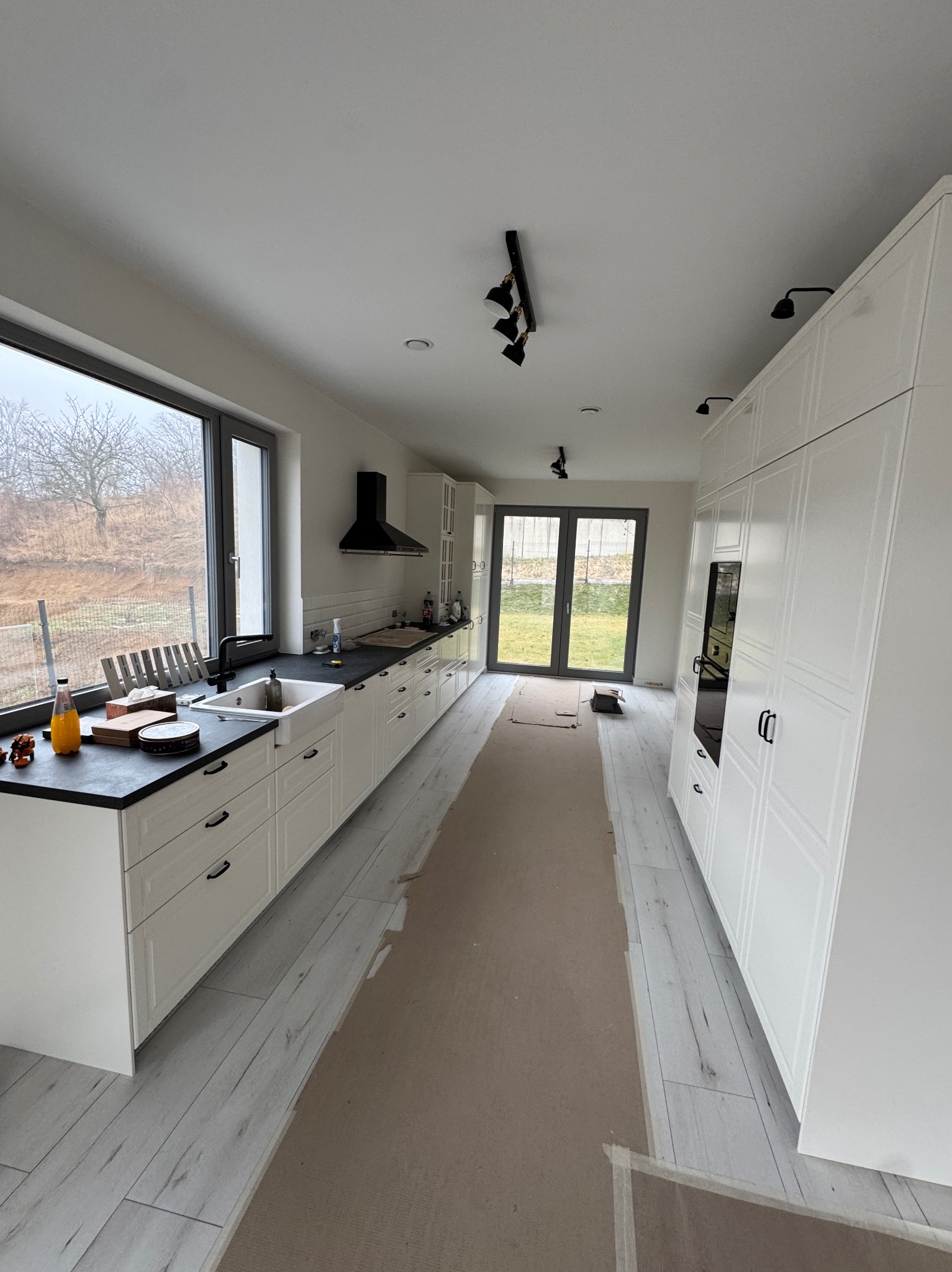 Jasna kuchnia z białymi szafkami i czarnym blatem, widok na okno i drzwi balkonowe. Podłoga wyłożona panelami imitującymi drewno. Oświetlenie punktowe.