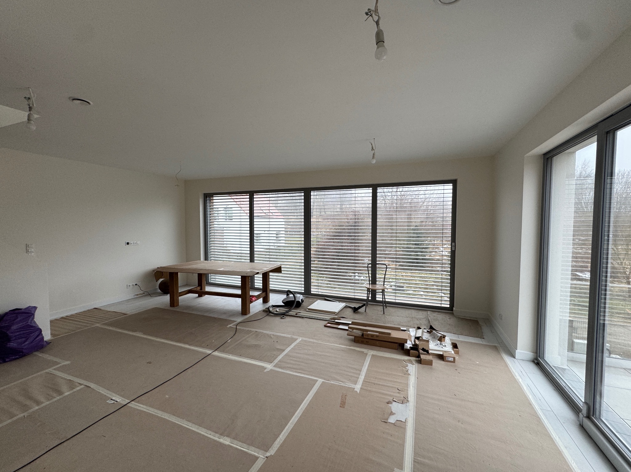 Przygotowanie do remontu: zabezpieczona podłoga kartonem, stół roboczy, okna z żaluzjami. Widoczne narzędzia i materiały budowlane. Wnętrze w trakcie prac wykończeniowych.