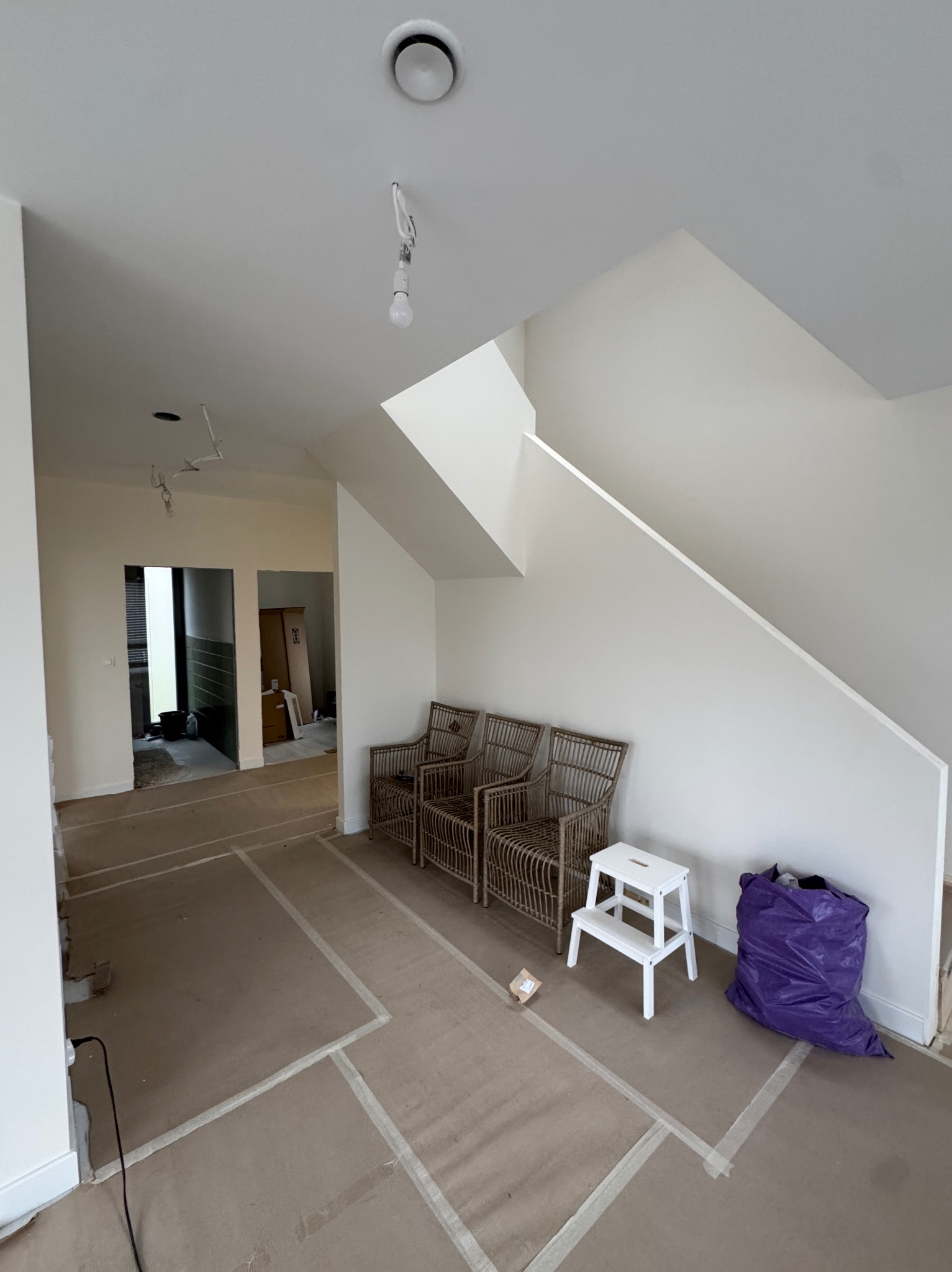 Jasne wnętrze w trakcie prac wykończeniowych: świeżo pomalowane ściany, zabezpieczona podłoga, wiklinowe fotele i schody w tle. Widoczny worek na śmieci i drabinka.