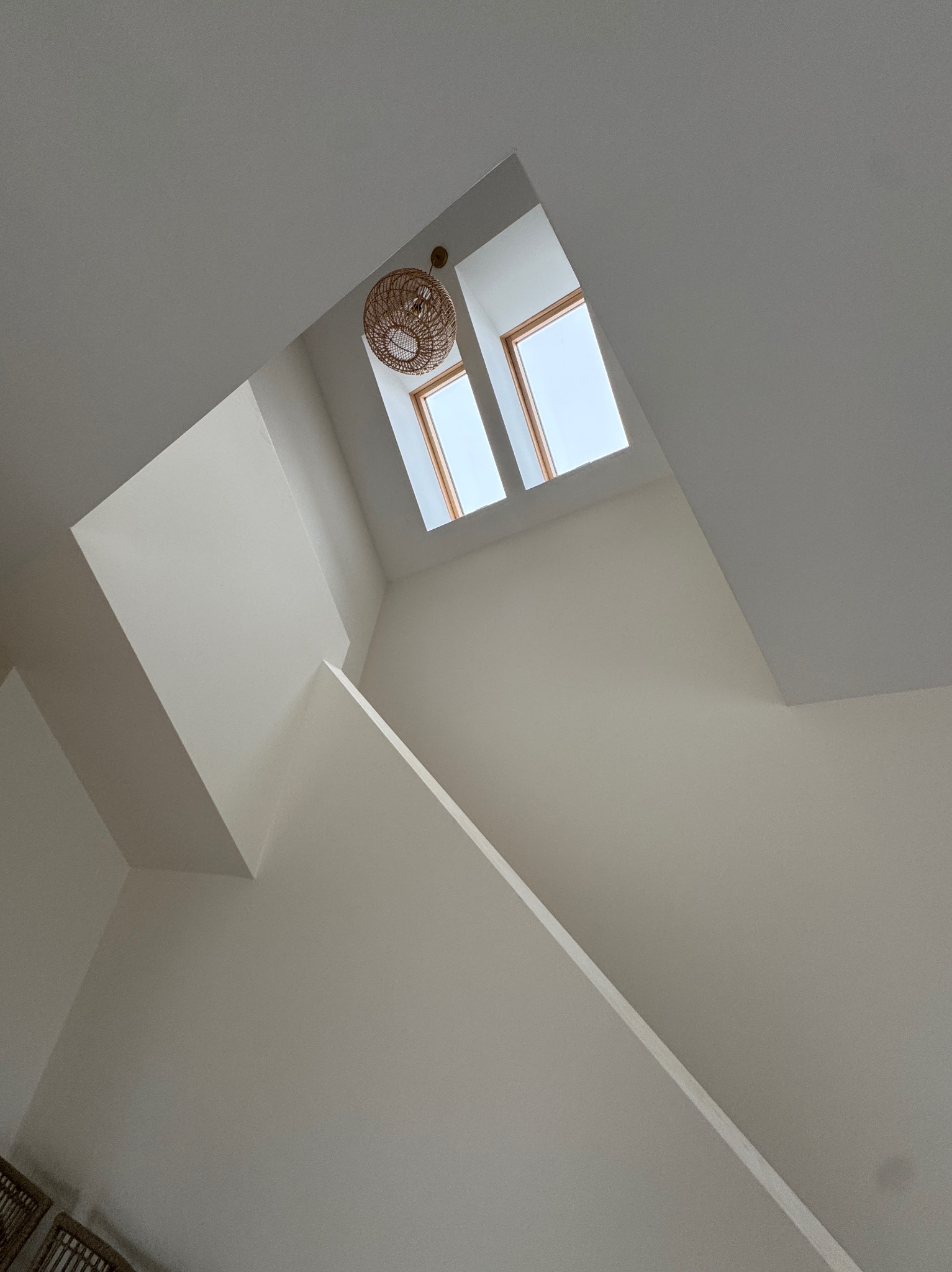 Białe ściany i sufit po malowaniu, widok z dołu na okna dachowe i lampę z wikliny. Geometryczne kształty i minimalistyczny design.