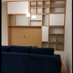ASGARD - MAREK BIERZOŃSKI - Drewniany regał z białymi elementami nad granatową sofą, oświetlony wiszącymi lampami. Widok na fragment salonu po montażu mebli.