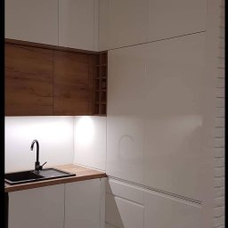 ASGARD - MAREK BIERZOŃSKI - Nowoczesna kuchnia w bieli i drewnie: lakierowane fronty, drewniane szafki wiszące z półkami na wino, czarny zlew i bateria, oświetlenie LED podszafkowe.