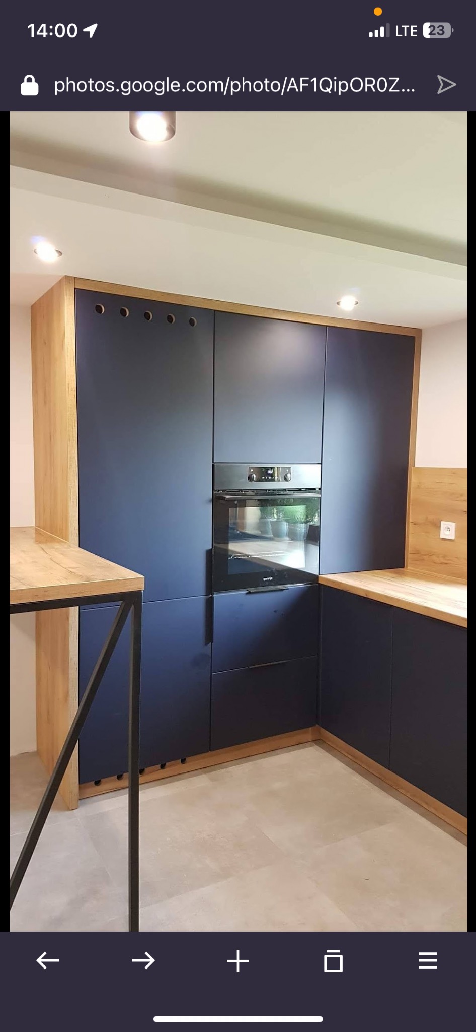 Nowoczesna kuchnia z ciemnoniebieskimi szafkami i drewnianymi akcentami. Widoczny piekarnik w zabudowie i fragment blatu. Oświetlenie punktowe.