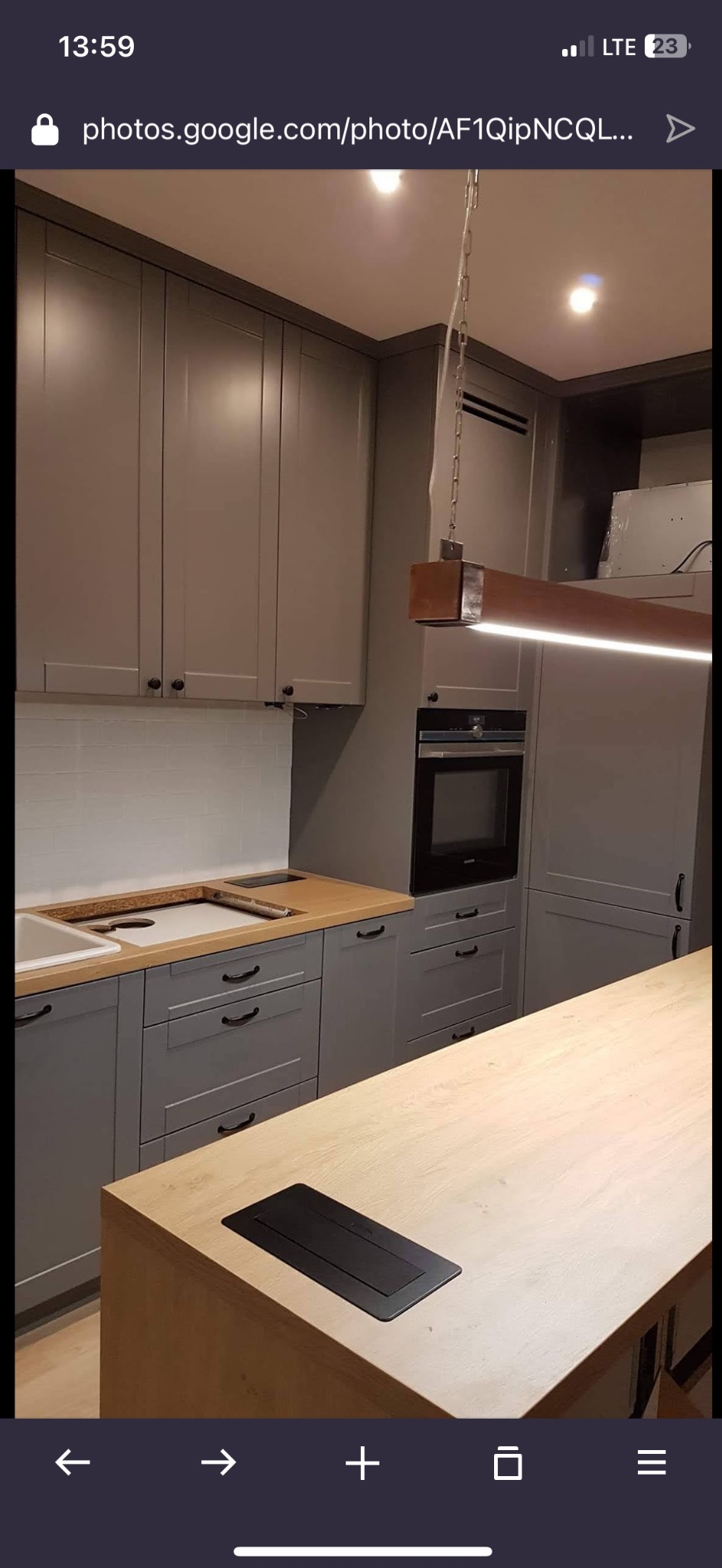 Szara zabudowa kuchenna z drewnianym blatem i wyspą. Widoczna biała glazura nad blatem i nowoczesna lampa wisząca. Ujęcie z perspektywy wnętrza kuchni.