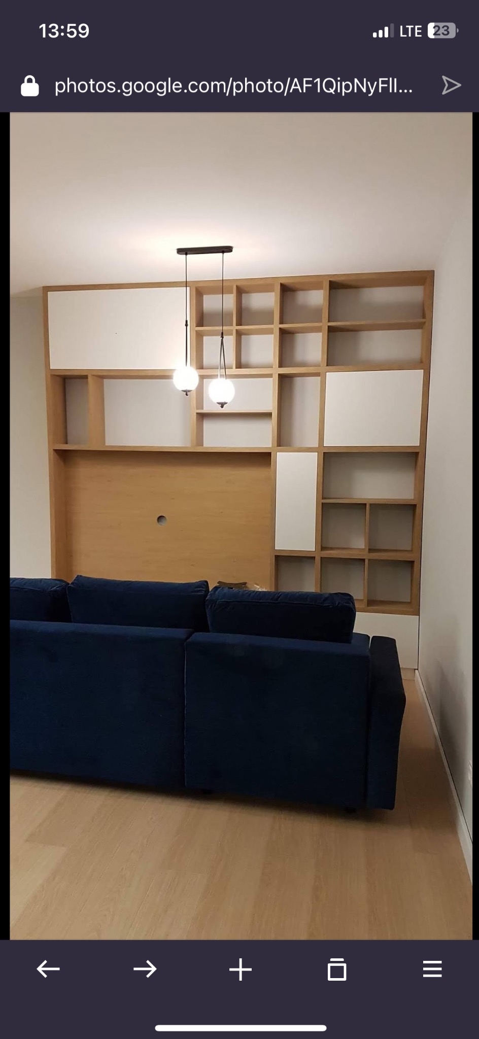 Drewniany regał z białymi elementami nad granatową sofą, oświetlony wiszącymi lampami. Widok na fragment salonu po montażu mebli.