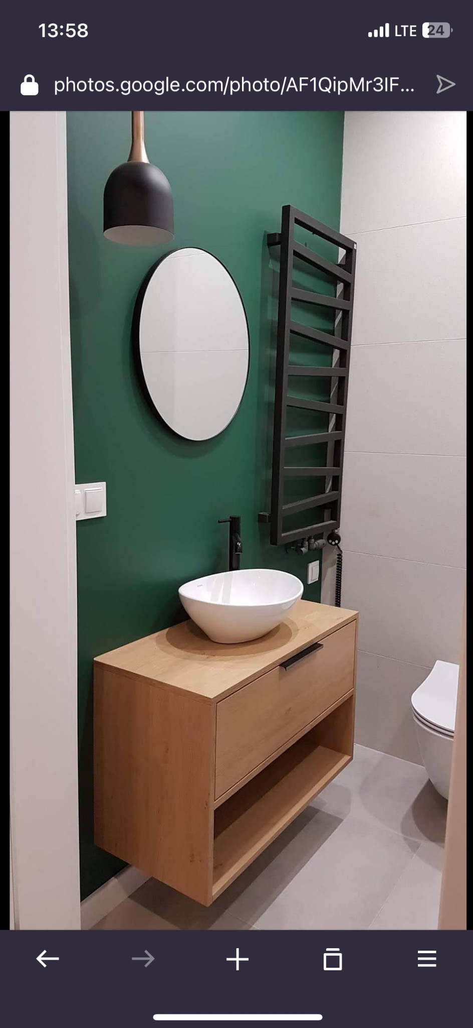 Nowoczesna łazienka z drewnianą szafką pod umywalkę, okrągłym lustrem i grzejnikiem drabinkowym na tle butelkowej zieleni. Minimalistyczny design, jasne płytki podłogowe.