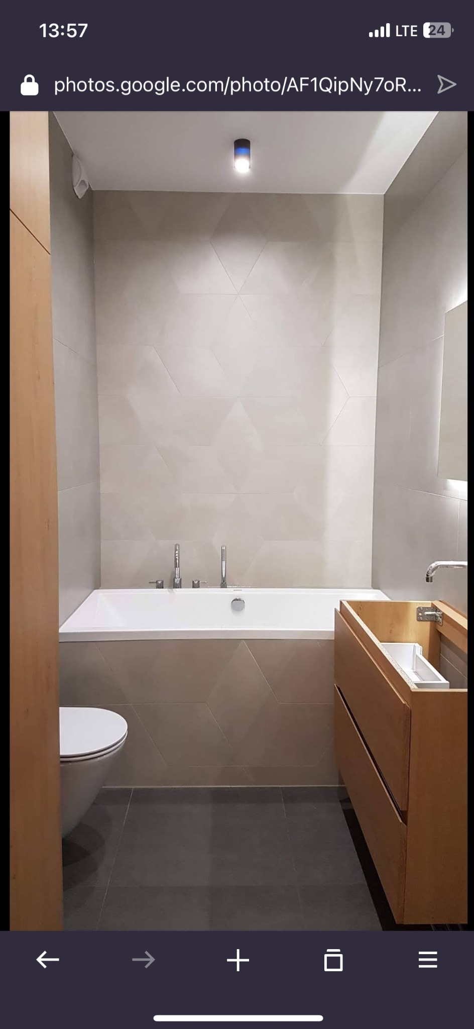 Nowoczesna łazienka z wanną obudowaną płytkami w geometryczny wzór, drewnianą szafką pod umywalką i szarą podłogą. Minimalistyczny design, stonowana kolorystyka.