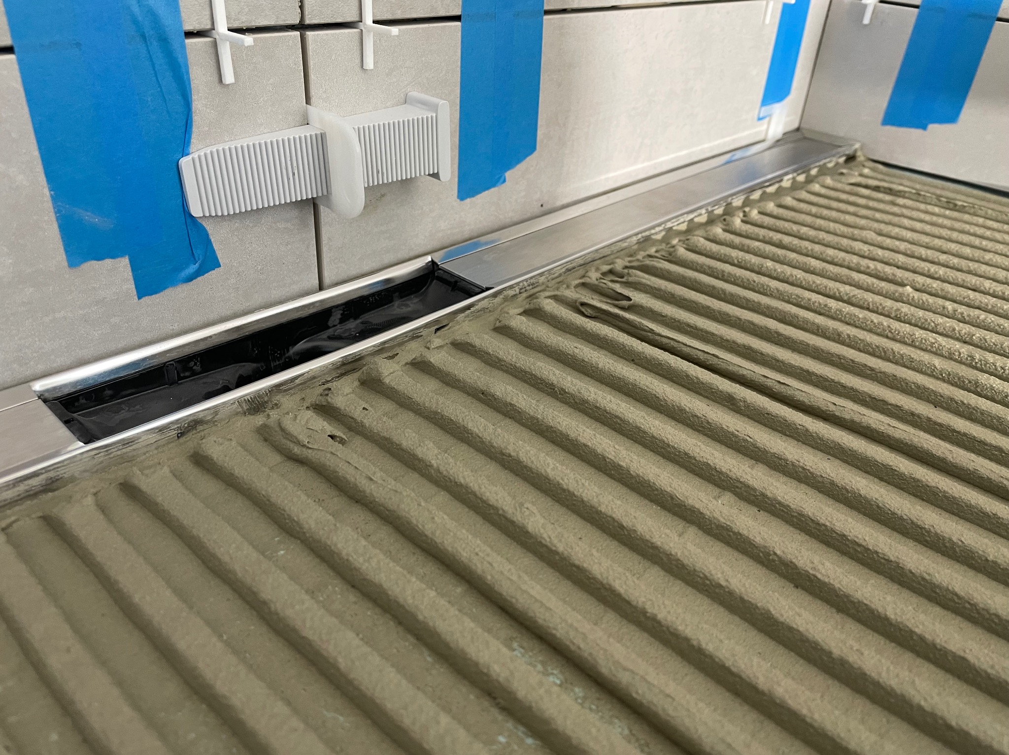 Ułożone płytki ścienne z niebieską taśmą i listwą odpływową. Na podłodze rozprowadzony klej z wyżłobieniami. Widoczne narzędzia glazurnicze.