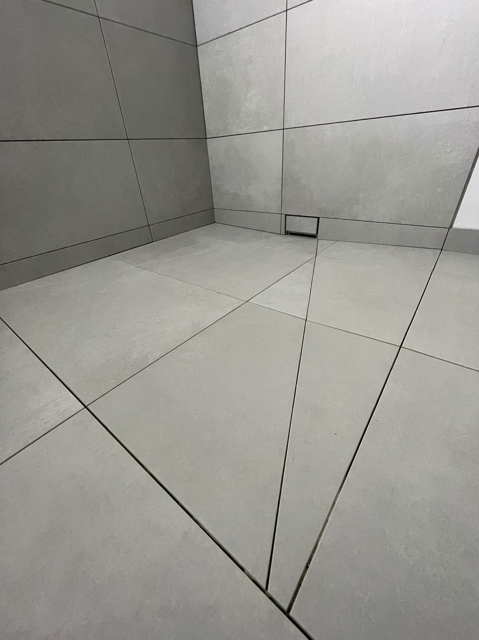 Minimalistyczna łazienka z szarymi, matowymi płytkami ułożonymi w kwadraty, z odpływem liniowym w rogu. Widok z góry podkreśla precyzję wykonania.
