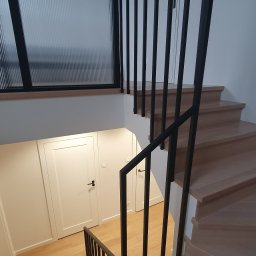 Norbert Turliński - Drewniane schody z czarną, minimalistyczną balustradą, widok z góry. W tle okno z ryflowaną szybą i białe drzwi. Jasne wnętrze, nowoczesny design.