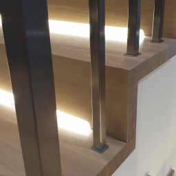 Norbert Turliński - Nowoczesne schody z drewnianymi stopniami i metalowymi słupkami balustrady, podświetlone od spodu, z bliska, ukazujące detale wykończenia i minimalistyczny design.