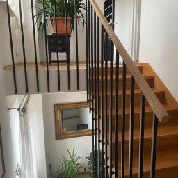 Norbert Turliński - Drewniane schody z czarną, metalową balustradą i drewnianą poręczą. Widok z góry, z elementami wystroju wnętrza w tle: lustro w złotej ramie, rośliny doniczkowe.