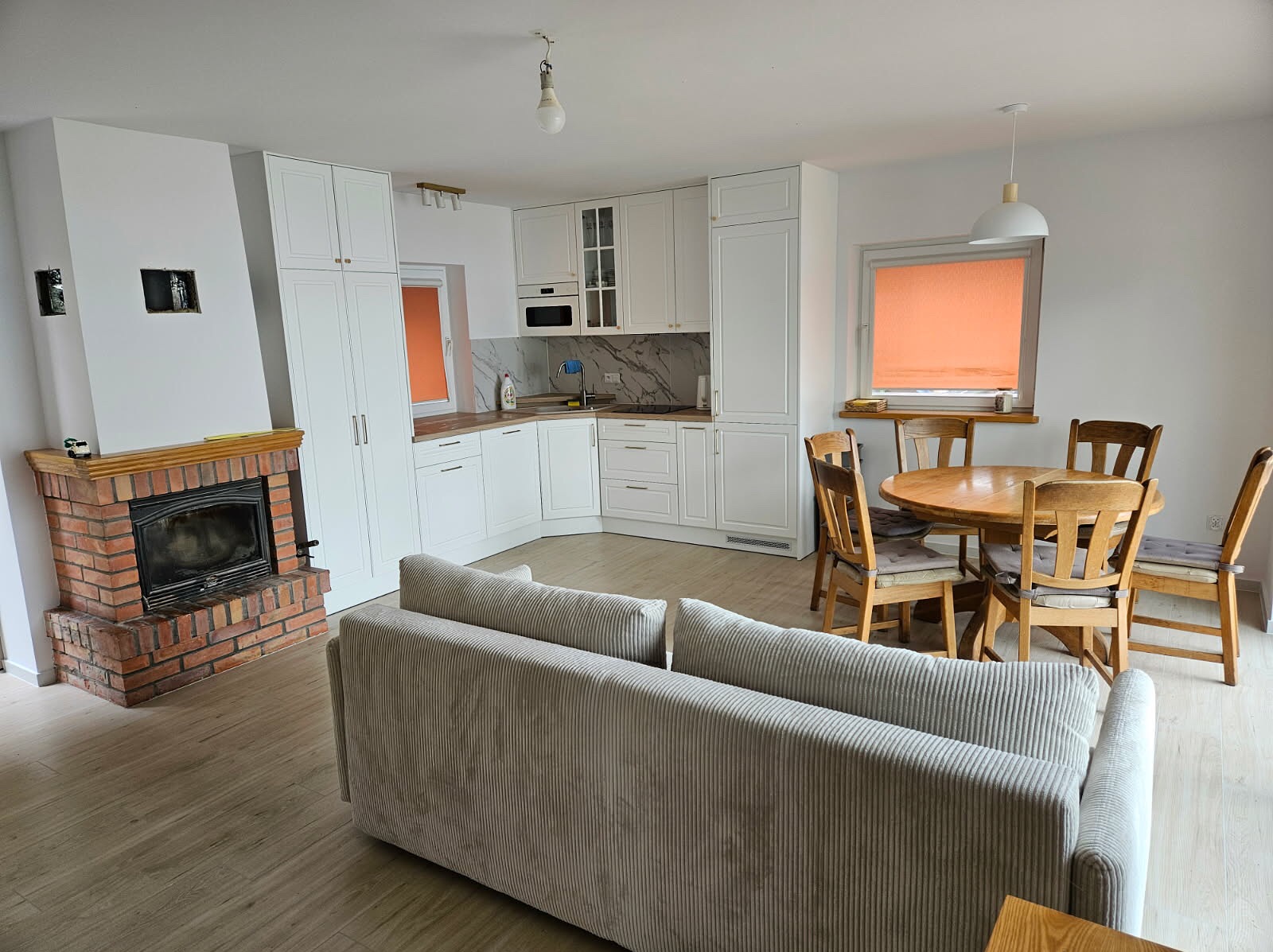 Jasne wnętrze: biała kuchnia z marmurowym blatem, ceglany kominek, drewniany stół z krzesłami i szara sofa. Całość w neutralnej kolorystyce, oświetlona naturalnym światłem.