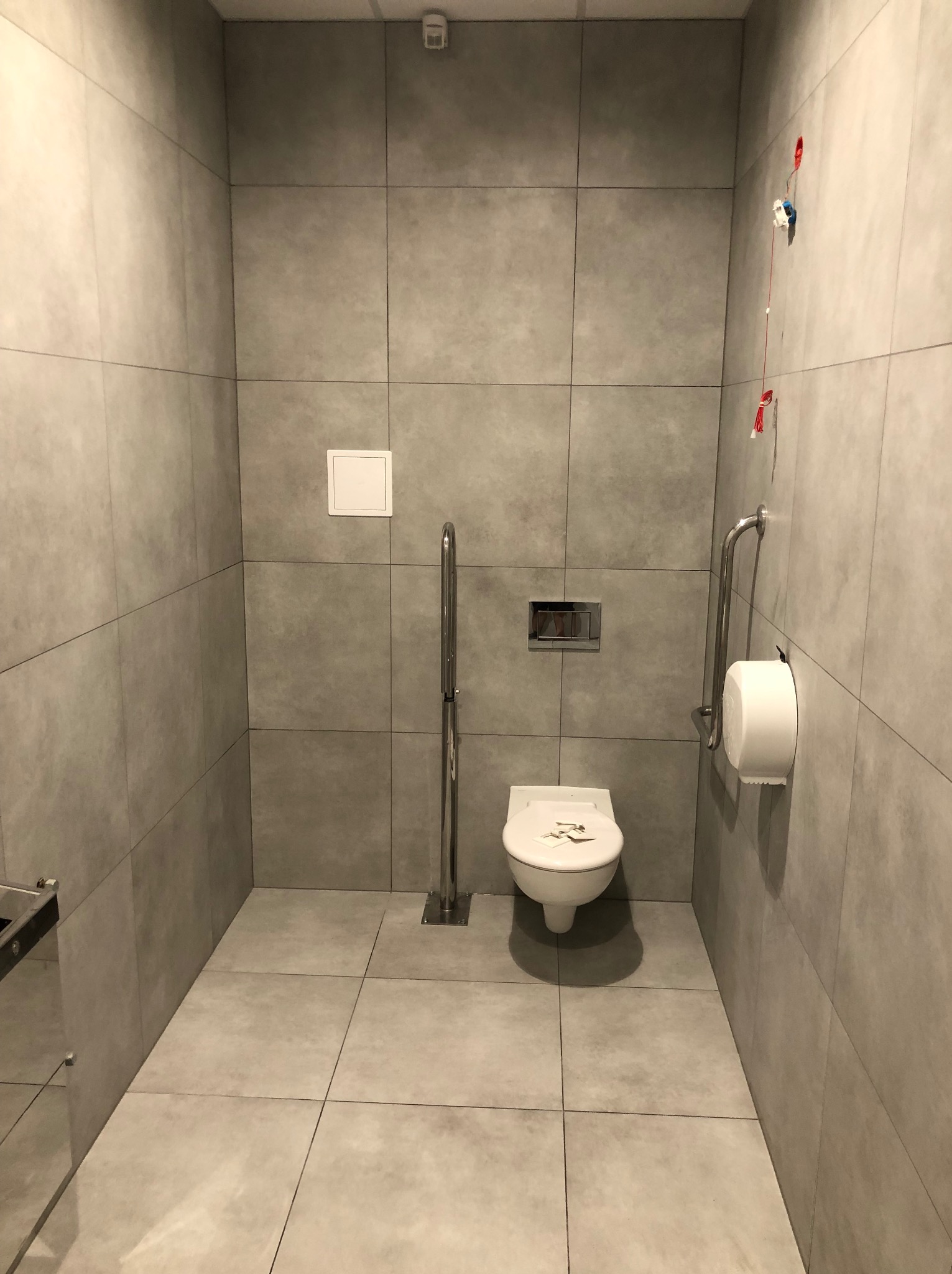 Wykończona łazienka w szarościach z toaletą dla niepełnosprawnych, uchwytami i dozownikiem papieru. Widoczne płytki wielkoformatowe i podwieszana toaleta.