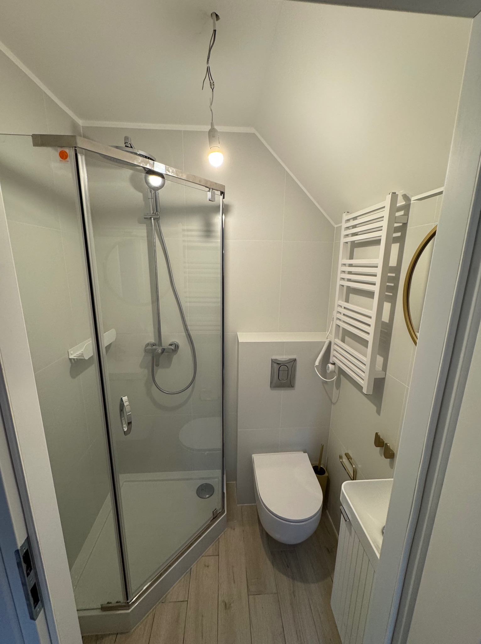 Jasna łazienka na poddaszu z kabiną prysznicową, toaletą, umywalką i grzejnikiem drabinkowym. Uwagę zwraca skośny sufit i drewnopodobna podłoga.