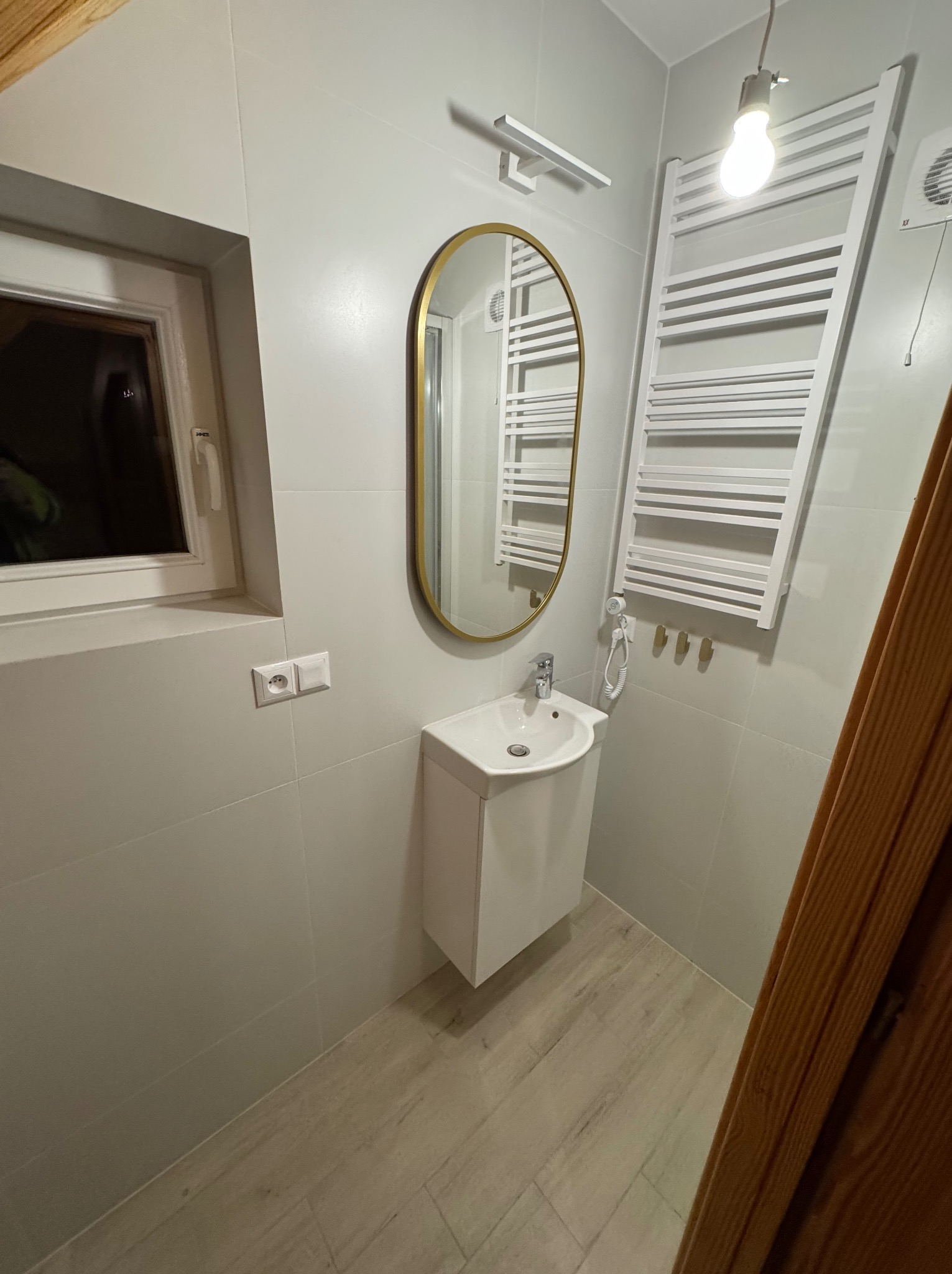 Jasna łazienka po remoncie: owalne lustro w złotej ramie nad białą umywalką z szafką, grzejnik drabinkowy, okno i drewniane drzwi. Minimalistyczny design.