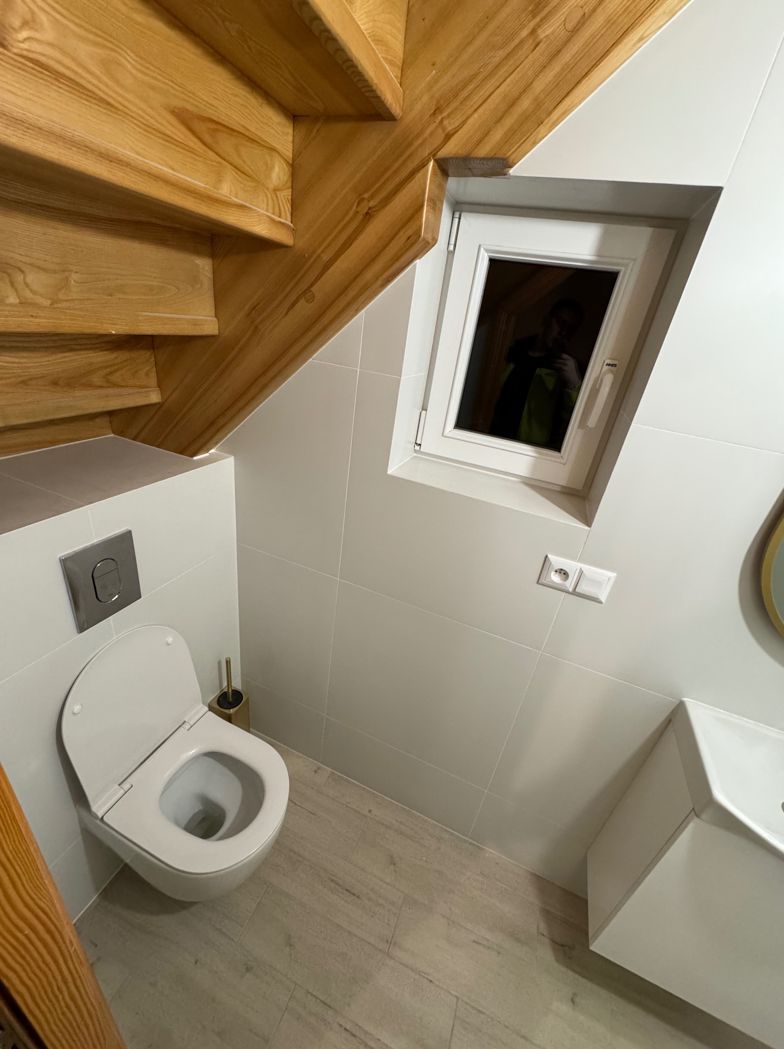 Nowoczesna łazienka na poddaszu z drewnianymi schodami, białymi płytkami i wiszącą toaletą. Okno dachowe wpuszcza światło. Minimalistyczny design.