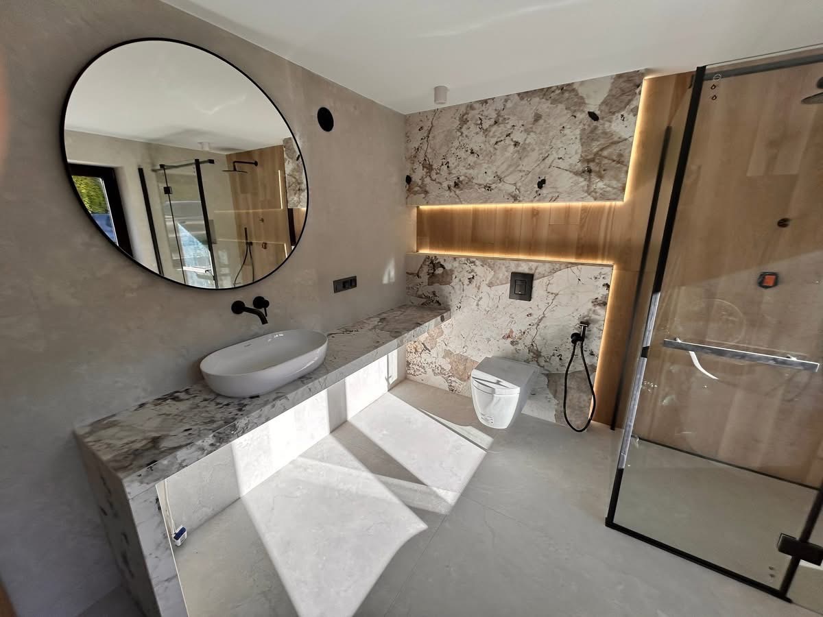 Nowoczesna łazienka z owalnym lustrem, marmurowym blatem i ścianą akcentową. Kabina prysznicowa ze szklanymi drzwiami i drewnianym wykończeniem. Minimalistyczny design.