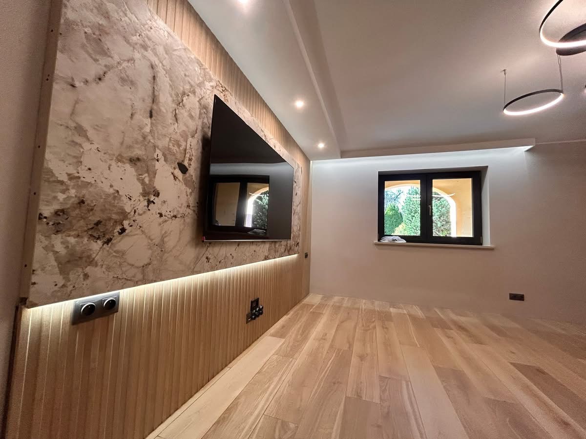 Eleganckie wnętrze: ściana z efektem marmuru, drewniane panele z oświetleniem LED, parkiet, okno z widokiem na zieleń i nowoczesne lampy sufitowe.