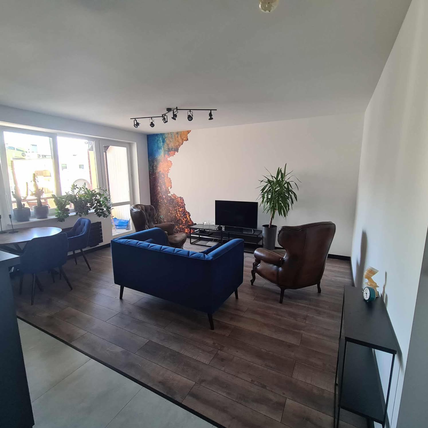 Salon po wykończeniu: drewnopodobna podłoga, niebieska sofa, skórzany fotel, ściana z abstrakcyjnym malunkiem, oświetlenie punktowe i rośliny doniczkowe.